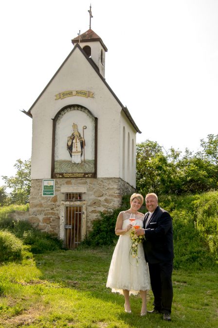 hochzeitsfoto das brautpaar steht vor der urbanuskapelle in traiskirchen und hält weingläser in der hand